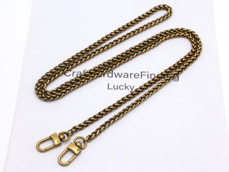 K&ouml;nnte beinhalten: Ein antiker, messingfarbener Kettenriemen mit Karabinerhaken. Die Kette hat ein gewebtes Design und wird auf einer wei&szlig;en Oberfl&auml;che pr&auml;sentiert. Der Text "Craft Hardware Finding Lucky" ist im Hintergrund sichtbar.
