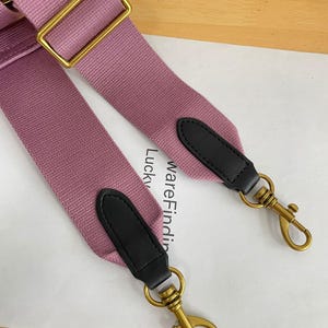Pode incluir: Uma alça de bolsa ajustável rosa empoeirado com ferragens de metal dourado. A alça tem detalhes em couro preto nas extremidades com fechos giratórios dourados. A alça é feita de tecido.