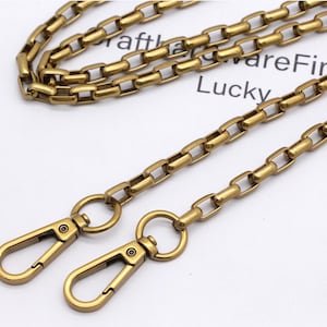 K&ouml;nnte beinhalten: Eine goldfarbene Metallkette mit rechteckigen Gliedern und zwei passenden Verschlussverschl&uuml;ssen. Die Kette liegt auf einer wei&szlig;en Oberfl&auml;che mit den W&ouml;rtern "craft" und "Lucky".