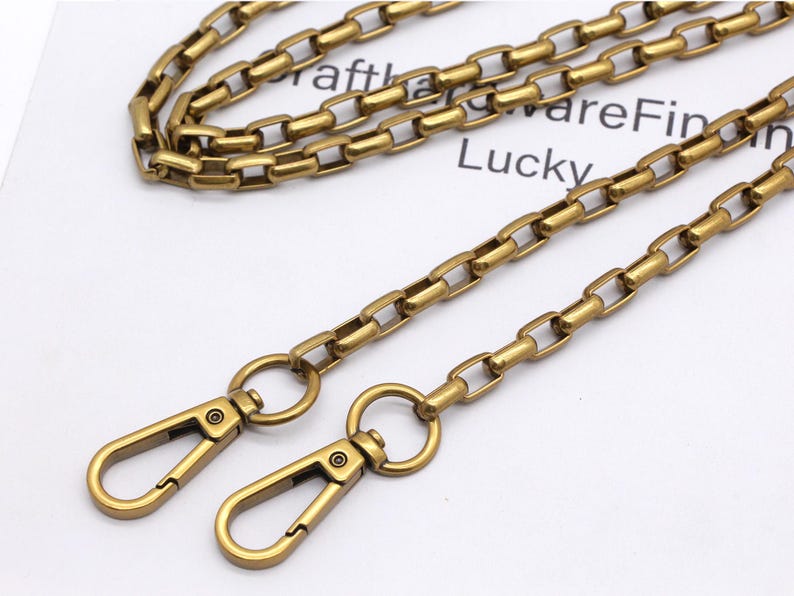 K&ouml;nnte beinhalten: Eine goldfarbene Metallkette mit rechteckigen Gliedern und zwei passenden Verschlussverschl&uuml;ssen. Die Kette liegt auf einer wei&szlig;en Oberfl&auml;che mit den W&ouml;rtern "craft" und "Lucky".