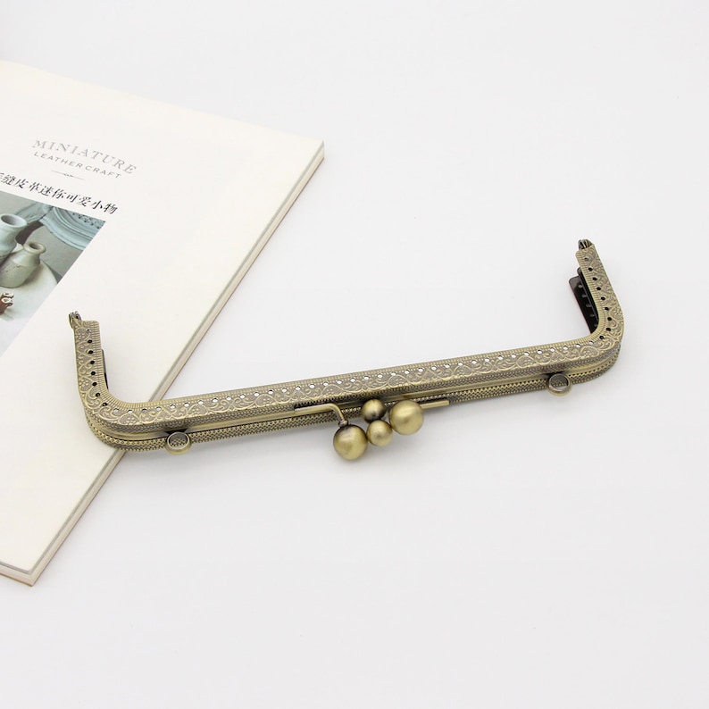 May include: A gold-toned metal purse frame with an ornate design and three decorative balls hanging from the center.
