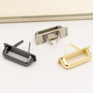 Könnte beinhalten: Drei Metalltaschengriffe in verschiedenen Ausführungen: Schwarz, Silber und Gold. Jeder Griff ist rechteckig mit abgerundeten Kanten und einer kleinen Schlaufe an jeder Seite.