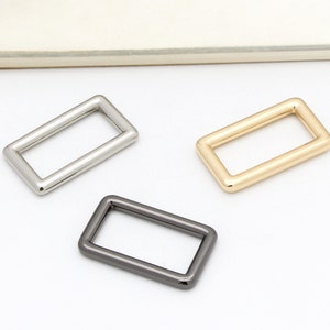 May include: Three metal D-rings in different finishes: silver, gold, and black. The rings are rectangular and have a smooth, polished surface.