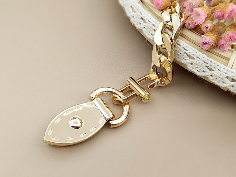 May include: Gold-toned metal clasp with a decorative oval-shaped piece. The clasp has a textured surface and a small, round, clear stone.
