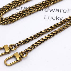 K&ouml;nnte beinhalten: Antik-messingfarbene Kette mit zwei passenden Verschl&uuml;ssen. Die Kette ist in einem gewebten Design gehalten, und die Verschl&uuml;sse sind im einfachen Hakenstil gehalten. Die Kette und die Verschl&uuml;sse befinden sich auf einer wei&szlig;en Oberfl&auml;che mit den W&ouml;rtern "Craft Hardware Fin" und "Lucky".