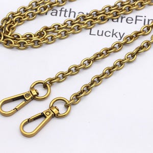 K&ouml;nnte beinhalten: Antik goldfarbene Kette mit zwei Karabinerhaken. Die Kette besteht aus kleinen, abgerundeten Gliedern. Die Verschl&uuml;sse sind mit einem kleinen Ring an der Kette befestigt. Das Wort "Lucky" ist im Hintergrund sichtbar.