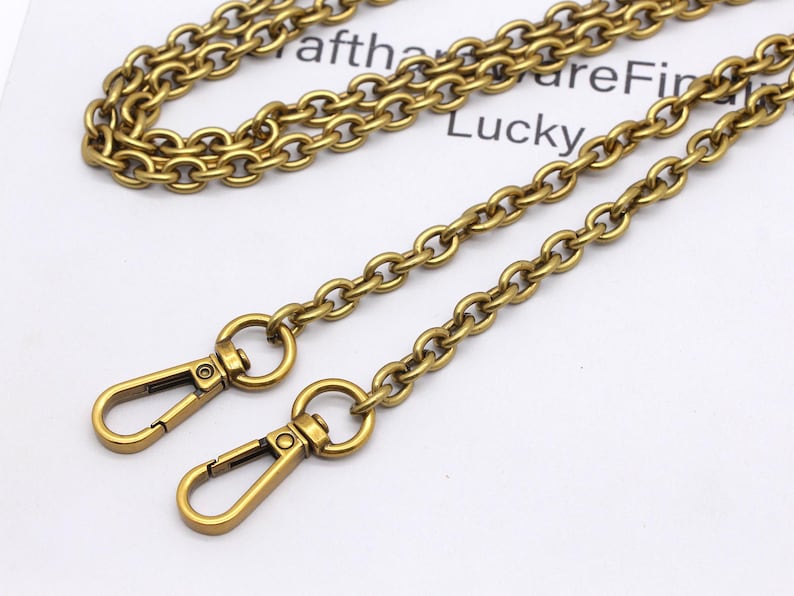 K&ouml;nnte beinhalten: Antik goldfarbene Kette mit zwei Karabinerhaken. Die Kette besteht aus kleinen, abgerundeten Gliedern. Die Verschl&uuml;sse sind mit einem kleinen Ring an der Kette befestigt. Das Wort "Lucky" ist im Hintergrund sichtbar.