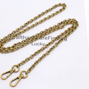 K&ouml;nnte beinhalten: Eine goldfarbene Kette mit zwei Karabinerhaken. Die Kette besteht aus ovalen Gliedern und liegt auf einer wei&szlig;en Oberfl&auml;che. Der Text "afthareFinding Lucky" ist teilweise im Hintergrund sichtbar.