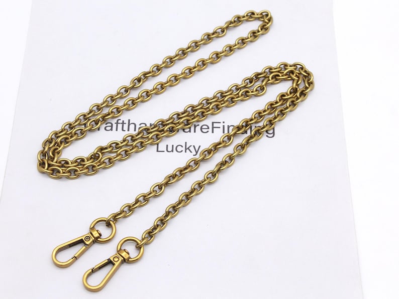 K&ouml;nnte beinhalten: Eine goldfarbene Kette mit zwei Karabinerhaken. Die Kette besteht aus ovalen Gliedern und liegt auf einer wei&szlig;en Oberfl&auml;che. Der Text "afthareFinding Lucky" ist teilweise im Hintergrund sichtbar.