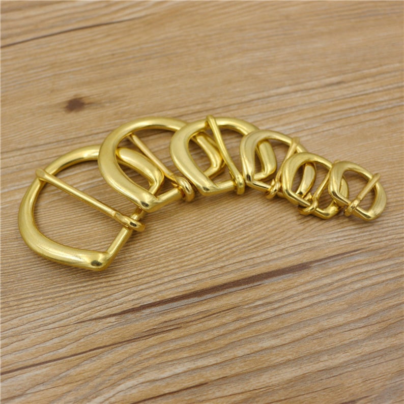 Puede incluir: Un juego de seis hebillas de cintur&oacute;n de metal dorado de diferentes tama&ntilde;os. Las hebillas est&aacute;n dispuestas en semic&iacute;rculo, con la hebilla m&aacute;s grande en la parte superior y la m&aacute;s peque&ntilde;a en la parte inferior.