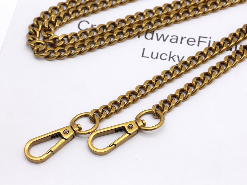 K&ouml;nnte beinhalten: Nahaufnahme einer goldfarbenen Kette mit zwei Karabinerverschl&uuml;ssen. Die Kette besteht aus ineinandergreifenden Gliedern und hat ein Vintage-Aussehen. Im Hintergrund ist der Text "HardwareFi Lucky" zu sehen.