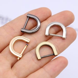 May include: Four D-ring metal hardware pieces in various finishes: gunmetal, silver, gold, and antique brass. These half-circle rings have a bar across the straight edge, used for attaching straps or other accessories. Ideal for crafting and leatherwork.
