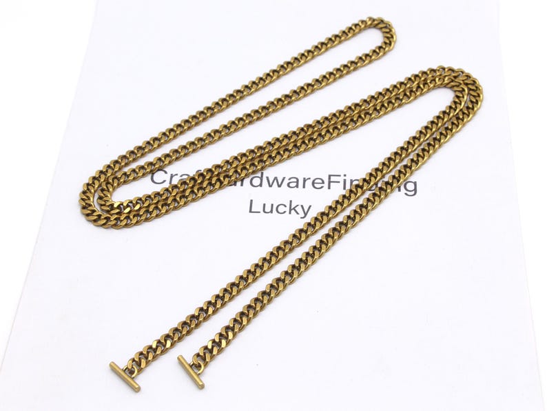 K&ouml;nnte beinhalten: Goldfarbene Panzerketten-Halskette mit T-Bar-Verschluss. Die Kette wird auf einer wei&szlig;en Oberfl&auml;che mit dem Text "Craft Hardware Finding Lucky" pr&auml;sentiert. Die Halskette ist aus Metall.