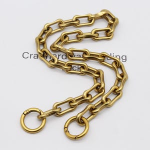 May include: A gold-colored chain with elongated links and two circular clasps. The chain is arranged in a curved pattern, and the text "Cra...ing" is partially visible in the background. This chain is likely for a bag or accessory.