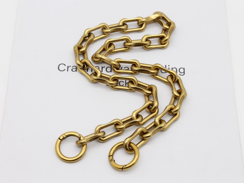 May include: A gold-colored chain with elongated links and two circular clasps. The chain is arranged in a curved pattern, and the text "Cra...ing" is partially visible in the background. This chain is likely for a bag or accessory.