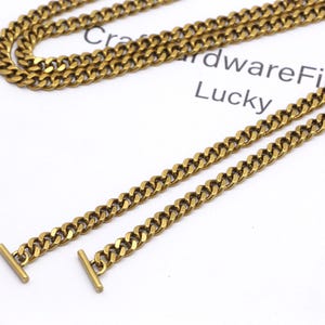 K&ouml;nnte beinhalten: Nahaufnahme von goldfarbenen Halsketten mit Curb-Link-Design. Eine Halskette ist gefaltet, zwei weitere sind gerade ausgelegt. Jede Kette hat einen T-Bar-Verschluss. Der Text "CraftwareFind Lucky" ist im Hintergrund sichtbar.