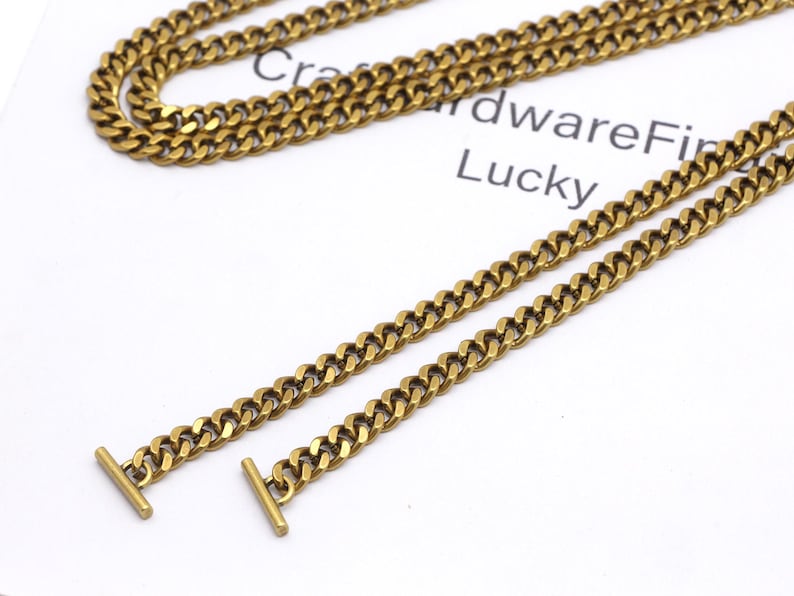 K&ouml;nnte beinhalten: Nahaufnahme von goldfarbenen Halsketten mit Curb-Link-Design. Eine Halskette ist gefaltet, zwei weitere sind gerade ausgelegt. Jede Kette hat einen T-Bar-Verschluss. Der Text "CraftwareFind Lucky" ist im Hintergrund sichtbar.