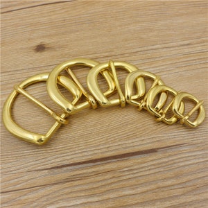 Puede incluir: Un juego de seis lazos de hebilla de cintur&oacute;n de metal dorado de diferentes tama&ntilde;os. Los lazos est&aacute;n dispuestos en semic&iacute;rculo sobre una superficie de madera.