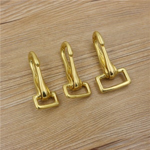 May include: Three gold-colored metal snap hooks with a swivel eye and a rectangular loop. The hooks are arranged on a light brown wooden surface. The hooks are likely used for attaching straps or other hardware.