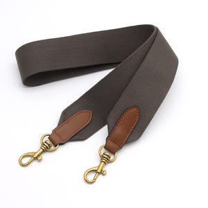 Pode incluir: Uma alça de bolsa cinza com detalhes em couro marrom e fechos de metal dourado. A alça é feita de um material tecido durável. Os fechos são projetados para serem presos a uma bolsa.