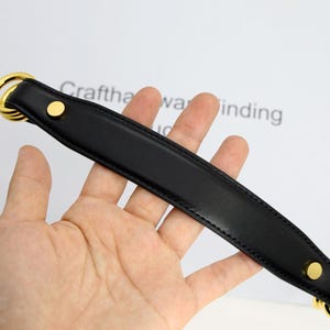 May include: Black leather bag handle with gold-coloured metal rings and rivets. The handle is held in a hand, with the text "Crafthawa winding" visible in the background. This accessory is designed for handbags and purses.