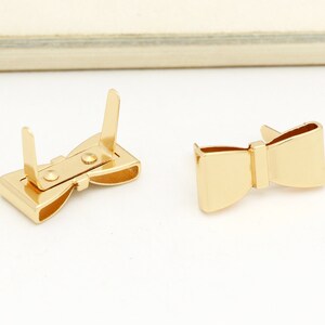 May include: Two gold-coloured metal bow-shaped clasps. One clasp is open, showing the mechanism. The other clasp is closed.
