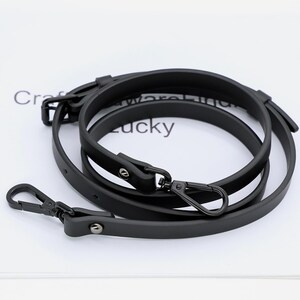 May include: Black leather bag strap with metal hardware. The strap is looped and features a swivel clasp and adjustable buckle. The word "Lucky" is partially visible in the background.