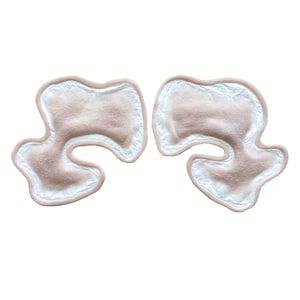 May include: Two flesh-toned, abstract-shaped pads with a white border. The pads have a soft, textured appearance and are set against a white background. The shapes are similar but not identical.