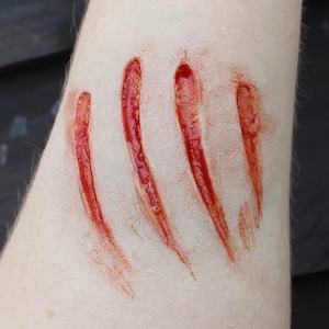 May include: Close-up of a forearm with four realistic-looking, deep red lacerations. The cuts are angled and appear fresh, with a glossy, wet texture, suggesting a special effects makeup application for a Halloween costume or theatrical production.
