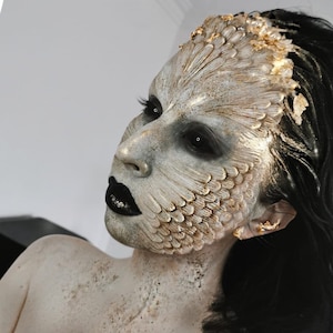 May include: A person with elaborate fantasy makeup featuring a textured, feathered design on the face, with black lipstick and dark eye makeup. The makeup is primarily beige and gold, with black hair.