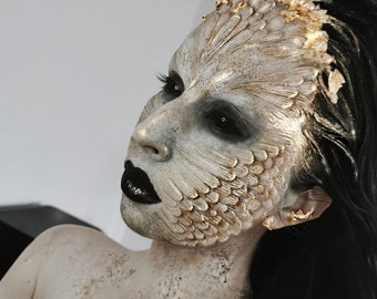 May include: A person with elaborate fantasy makeup featuring a textured, feathered design on the face, with black lipstick and dark eye makeup. The makeup is primarily beige and gold, with black hair.