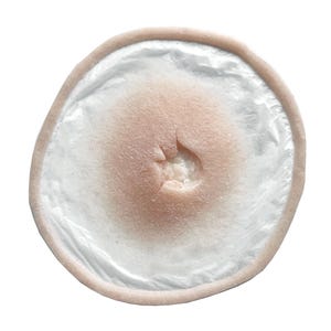 May include: A flesh-toned prosthetic wound with a central indentation and a raised edge. The wound is surrounded by a translucent, white material, creating a layered effect. The overall shape is circular.