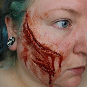 Könnte beinhalten: Nahaufnahme eines Gesichts mit einer großen, blutigen und realistisch aussehenden, falschen Wunde auf der Wange.