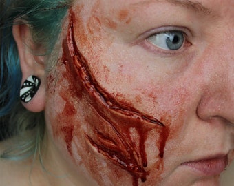 May include: A close-up of a person's face with a large, bloody, and realistic-looking fake wound on their cheek.