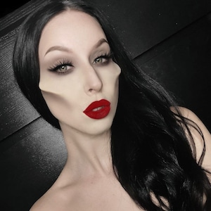 May include: A woman with long black hair and pale skin is wearing red lipstick. Her face is partially obscured by a white mask.