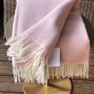 May include: A light pink blanket with cream-colored fringe is draped over a gold-colored stand. The blanket is folded, showcasing its soft texture. The background includes a wicker chair and wooden floor.