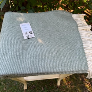Puede incluir: Una manta de color verde salvia con un patrón de tejido de espiga diagonal y flecos de color crema en un extremo. Una etiqueta blanca está adherida a la superficie con el texto 140x200 cm, indicando las dimensiones de la manta.