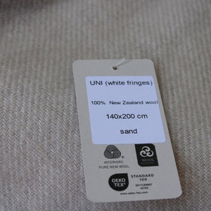 Op de afbeelding: Een zandkleurige wollen deken met witte franjes. Het label geeft aan dat het 100% Nieuw-Zeelandse wol is, met een afmeting van 140 x 200 cm. De tekst "UNI (white fringes)" en "sand" is ook aanwezig.