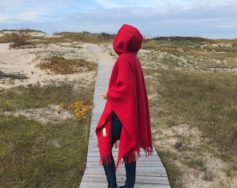 May include: A red hooded poncho with fringe trim. The poncho is worn by a person walking on a wooden path.