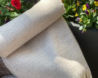 Può includere: Una morbida coperta di colore grigio chiaro con frange bianche. La coperta è piegata e appoggiata su una sedia.