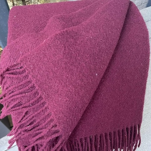 May include: A folded claret throw blanket with fringed edges. The blanket is made of a soft, textured material and is draped over a surface. The fringe is a darker shade of claret.