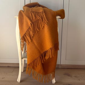 Op de afbeelding: Een effen mosterdgele plaid met een getextureerd, geweven uiterlijk. De deken is voorzien van lange, bijpassende franjes langs de randen, wat zorgt voor een zachte en gedrapeerde uitstraling terwijl deze over een licht houten meubelstuk hangt.