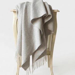 May include: A soft, light beige wool throw blanket with a textured, felted appearance and long, straight fringe detailing along the bottom edges. The blanket is draped over the back of a light-coloured wooden chair, showcasing its folds and drape.