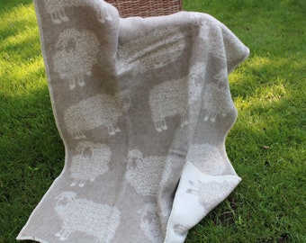 May include: A beige wool throw blanket with a white sheep pattern. The blanket is folded and draped over a wicker basket.