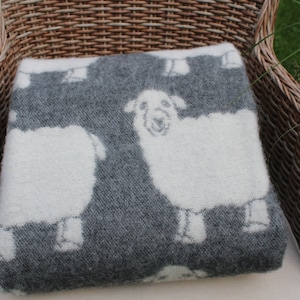 May include: A grey and white sheep patterned throw blanket is draped over a wicker chair. The blanket is made of soft, fluffy material and features a repeating pattern of white sheep on a grey background.
