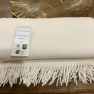 Op de afbeelding: Een witte wollen deken met witte franjes. De deken is dubbelgevouwen en heeft een label met de tekst "Lini (white fringe) 100% Pure New Wool 140x200 cm natur".