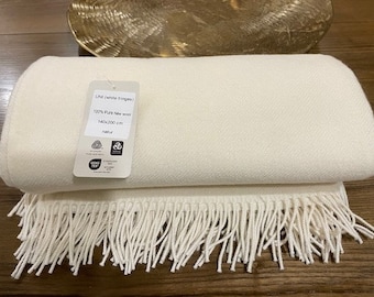 May include: A white wool throw blanket with white fringe. The blanket is folded in half and has a label that reads "Lini (white fringe) 100% Pure New Wool 140x200 cm natur".