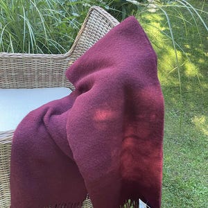 May include: A deep burgundy throw blanket with fringed edges, draped over a wicker chair. The blanket has a soft, textured appearance. The scene is set outdoors on a sunny day. The blanket is a rich shade of red.