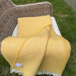 Puede incluir: Una manta amarilla con estampado de espiga y flecos blancos está sobre una silla de mimbre. La manta es de un color dorado cálido y parece suave y acogedora. La silla está sobre una superficie cubierta de hierba.
