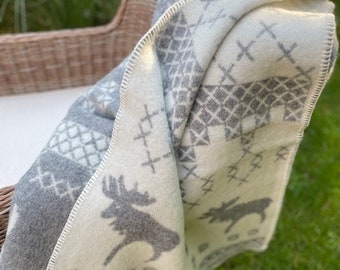 May include: A white and gray wool throw blanket with a Scandinavian-inspired pattern featuring reindeer and geometric designs. The blanket is folded and draped over a wicker chair.